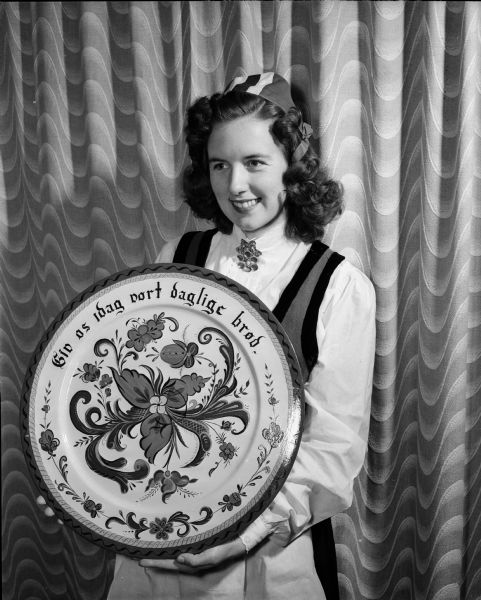 Mrs. Robert (Dorothy L.) Sirney, standing in front of a curtain and wearing a Norwegian costume. She is holding a wooden platter she has decorated with rosemaling, following the tradition of original designs. Mrs. Sirney is of Norwegian descent.