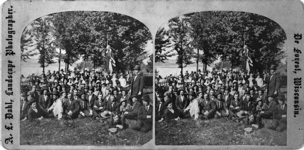 Group portrait of a large group of men, women and children, at a lake shore, possibly Maple Bluff on Lake Mendota near Madison. The Norwegian flag suggests that this may be Syttende Mai (May Celebrations).