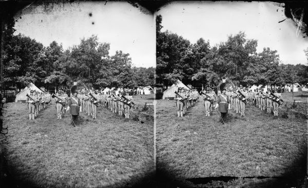 Quite possibly this is the "View of the Grand Music Band" listed in the "Centennial Views of the City of Madison, July 4th, 1876. Views of Chicago Light Guard" section of Dahl's 1877 "Catalogue of Stereoscopic Views." It appears to be a Marine Corps band encamped at Camp Randall or near Lake Monona in Madison in preparation for the Centennial Celebration on July 4, 1876. The men stand in ranks with their band instruments, their leader is in the foreground and tents are behind them.