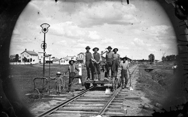 Group portrait of a railroad crew on and around a handcar. A man and a young boy stand on the left near a track switch. In the background is a variety store and other buildings. A small sign indicates track or mile 103.