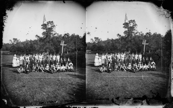 Large group at Syttende Mai celebration. Group posing with what may be the national and merchant flag of Norway flag. A number of people are holding croquet mallets. There is a U.S. flag flying on a flagpole in the background among trees.