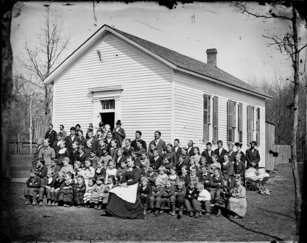 The first school house in DeForest is a frame, one-story building with shutters. The group posed in front consists of the woman teacher and her students. Behind them are standing men and women parents. A man and a young girl stand behind a baby carriage on the far right, with a Dalmation dog lying underneath. Louis Dahl is seated in the front right row with a cap on his knee.