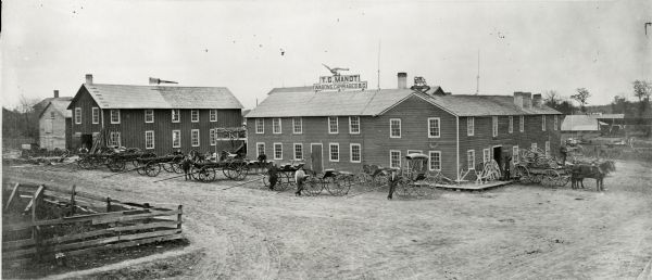 Slightly elevated exterior view of T.G. Mandt Wagons & Carriages. Several of the men are standing near or leaning on one of the many carriages displayed around two main buildings. A bell is mounted on the roof of the largest building.