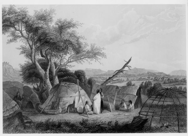 Indians standing and sitting near wigwams. Four wigwam frames are covered, with the one on the right uncovered, with a campfire inside. In the distance are tree-covered hills and bluffs.