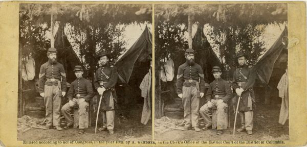 Stereograph of three U.S. Army soldiers in front of tents in camp under a framework of branches.