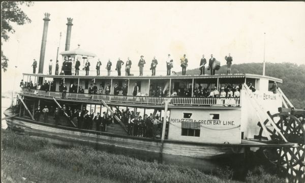 View from shoreline of a large crowd of people posing on the decks of a docked steamboat. There is a row of men standing along the top of the ship. On the far shoreline is a tree-lined hill or bluff. The words "City of Berlin" are painted along the back of the steamboat. On the side is painted: "Portage City E. Green Bay Line."