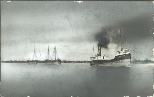 Photographic postcard of a steamboat and several other ships on serene water. There is a shoreline in the far background.