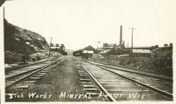 View down railroad tracks that lead to industrial buildings and a smokestack. A caption on the image reads, "Zink (Zinc) Works, Mineral Point, Wis."