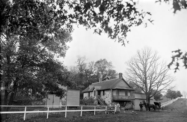 View of the Dawn Manor coach house, which shows a woman standing next to the fence and a man posing with a horse-drawn carriage.