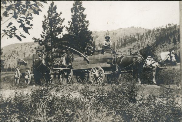 Three men pose on two wooden horse-drawn wagons. A bluff is in the far background.