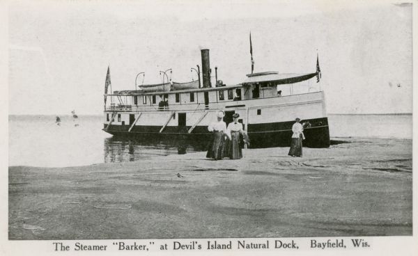 Three women stand on the shore in front of the steamer <i>Barker</i> at Devils Island natural dock.