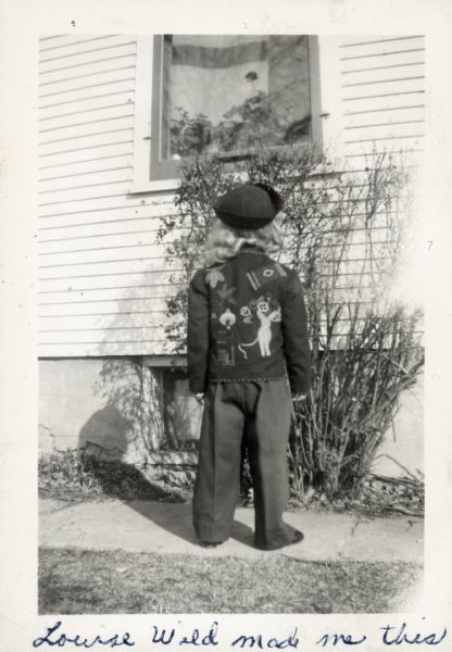 A back view of Linda Schiesser (b. 1944) posing outdoors wearing trousers, a hat, and a Mexican Tourist Jacket. Written at the bottom of the print: "Louise Wild made me this."