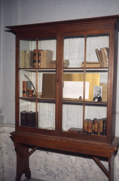 Bookcase in the second floor hallway outside the library of the State Historical Society of Wisconsin. The case contains 50 books that were part of the original collections of the Society. The case also holds a photograph of Lyman Draper and a brief interpretive paragraph.