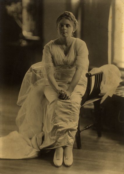 Full-length portrait of Dorothy Turner Main seated with her hands clasped in her lap. She wears a ankle-length dress and a necklace.