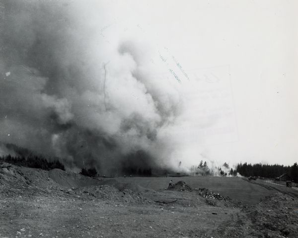 View across a field towards thick smoke billowing from a forest fire. People are in an open field in the distance. A road with trucks parked along it runs up to a small building on the right.