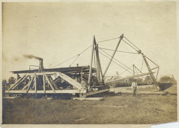 View of a walking dredge in a field. A man, a woman holding a child, and a young boy are on the dredge, and a woman perches on the projecting arm, and another man stands in the grass on the right. Buildings are in the far background.