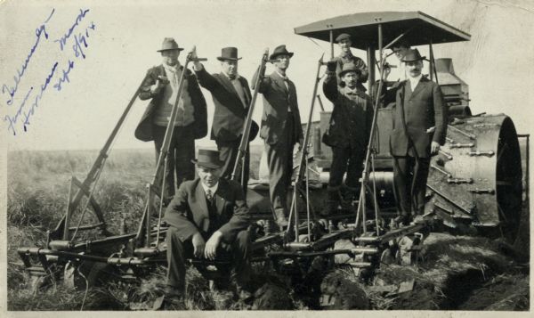Five men in hats and jackets, some holding the levers on a gang plow pulled by a Rumley tractor, pose for a group portrait. One man in work clothes stands on the tractor and another in a coat and hat with a cigar in his mouth sits on the plow in the foreground. The plow was likely part of the effort to drain Horicon Marsh for development.