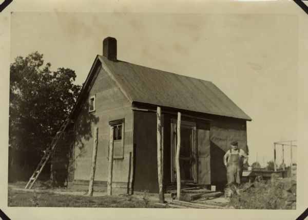 Man in coveralls standing outside a small house. A ladder leans against the side of the house on the left. There are four wooden posts in the foreground.