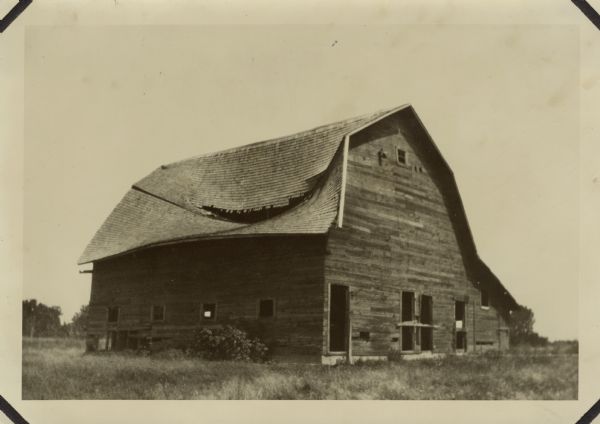 View of an abandoned barn in a field with long grass. The roof is caved in.