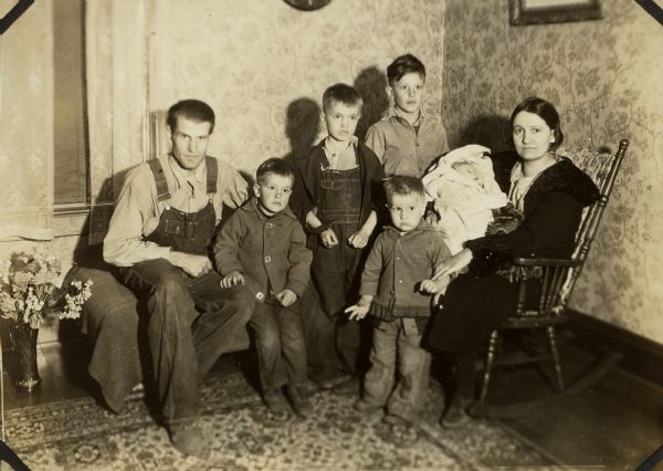 Brundage family in their new home after resettlement with assistance of United States Resettlement Administration. From left to right: Walter, Marvin (4), Bruce (7), Robert (11), Victor (2), Harold Jean (1 month), and Thelma. A vase of cut flowers sits on the floor at left. Thelma holds the baby while seated in a rocking chair.