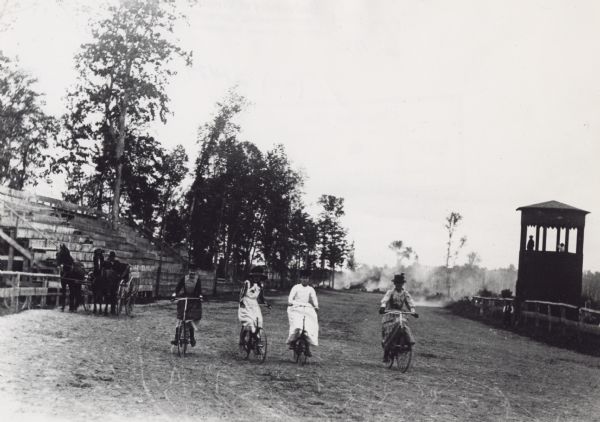 Four women in dresses and hats riding bicycles on a race track at the Langlade County Fair Grounds. Bleachers at left are empty, but there is a man in a carriage pulled by two horses in front of the stands. On the right two people are in the judge's stand and another man stands on the track below.