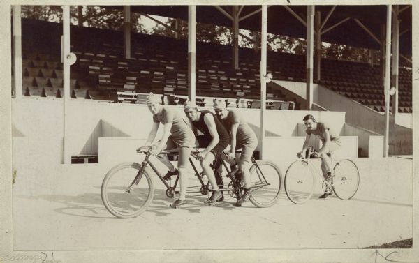 Three men (Charles Crane, unidentified man, and Stuart Harbridge) in racing gear on a triplet tandem bike in front of racer Al Bauman on the Racine Athletic Association's cement bicycle racing track.