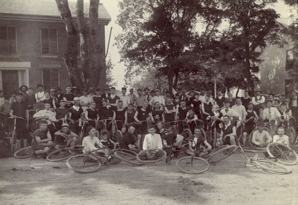 Portrait of a large group of mostly men in touring clothes posed outdoors with bicycles.