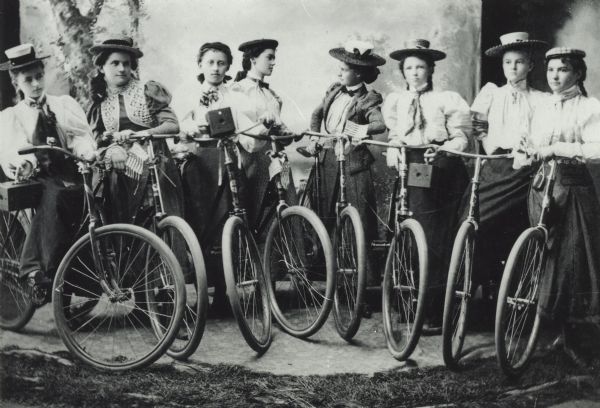 Studio portrait in front of a painted backdrop of eight young women, each standing and holding a bicycle. Three of the women also hold brownie cameras.