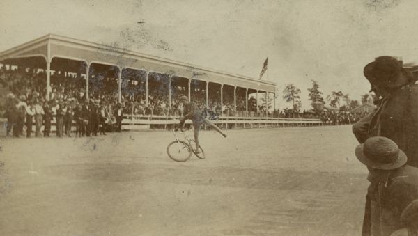 Lee Richardson performing stunts on a bicycle in front of a large crowd standing on the ground, as well as seated in bleachers at the Appleton County Fair and Driving Park during the League of American Wheelmen meet.