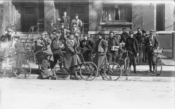 Informal portrait of a group of people gathered on the curb and sidewalk in front of a building. Many of the people have bicycles. There is a pennant behind a women in the center that reads in part: "Wauch" perhaps for "Waucheta."
