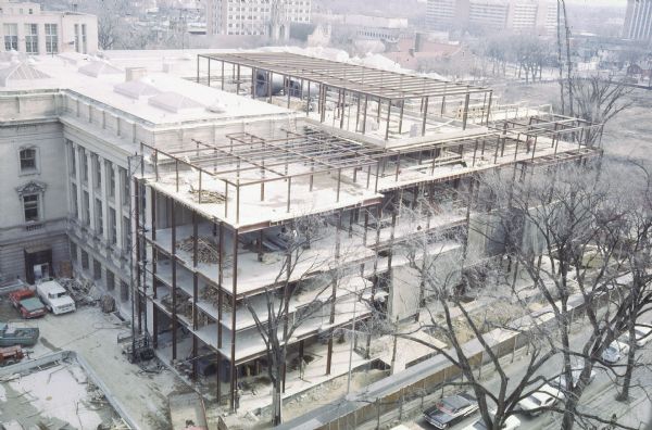 Elevated view of construction of the T-shaped addition on the Wisconsin Historical Society Headquarters Building, from Science Hall looking southeast. Vehicles are in the parking lot at left and along Park Street in the foreground. Memorial Library is in the background on the left.