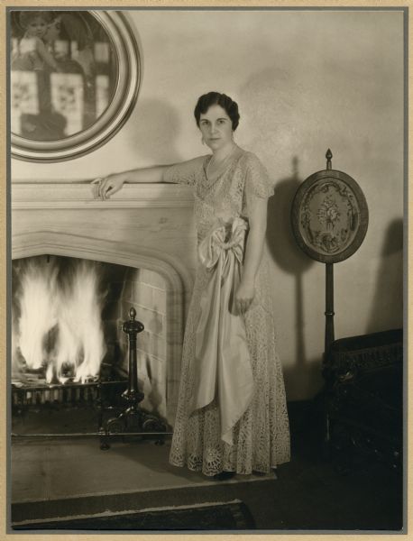 Formal full-length portrait of Dorothy Werner standing at a fireplace. She wears a dress that is now held by the Wisconsin Historical Society.