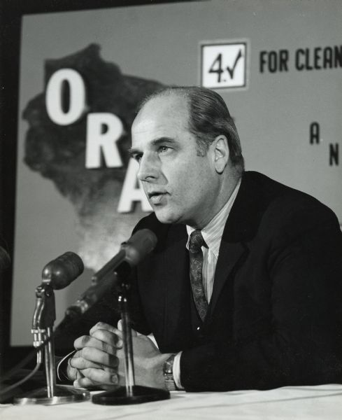 Governor Nelson seated and speaking into microphones about his Outdoor Recreation Action Program (ORAP), the 1961 proposal for Wisconsin land purchase and conservation. An ORAP sign is on the wall behind Nelson.