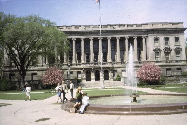 View of the State Historical Society of Wisconsin headquarters building from across Library Mall. A woman and child are sitting on the edge of the William Hagenah Memorial Fountain and other people are walking through the mall. Trees on both sides of the front stairs are flowering. The U.S. flag flies at center.