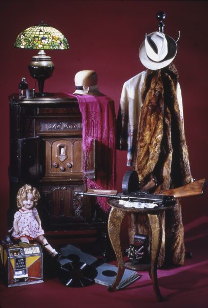 Still life collection of a number of items from The Wisconsin Historical Society Museum collections, including a fur coat, a slot machine, a Tiffany lamp, a doll, a radio, hats, a gun, phonograph records, and a table.