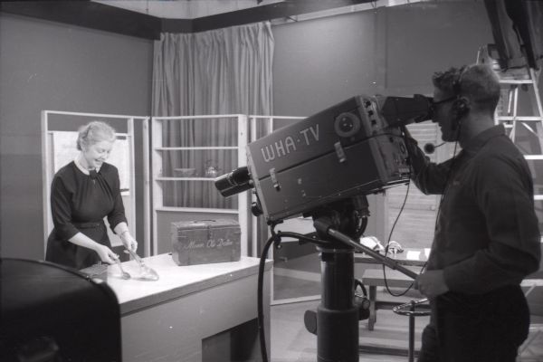 Doris Platt at the WHA-TV station displaying artifacts during the broadcast of <i>Scandinavian Backbone</i>, part of her series of 1959 television shows. Edward Crawley is the WHA cameraman. Another person is standing on a ladder in the background on the right.