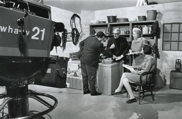 Doris Platt (standing) and three other people, possibly discussing the script of a program on WHA-TV. Artifacts line shelves behind the people in a country store setting. The WHA camera is in the left foreground. One man is smoking a pipe.