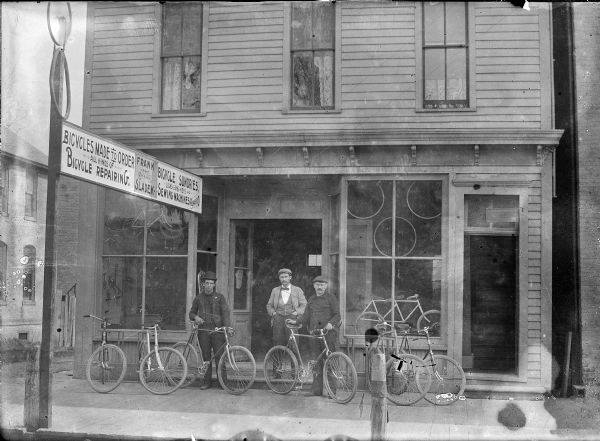 View from street of three men standing with bicycles in front of Frank Sladek's bicycle shop. A sign at left indicates that Sladek's shop custom built and repaired bicycles and also sold locks, guns, keys and repaired sewing machines.