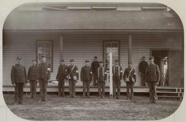 Outdoor group portrait of the Hospital Crew of the 1st Wisconsin Regiment. Two men wear canteens and bags. Others wear straps around their necks that were likely used to assist in carrying stretchers. The 3rd and 4th men from the left are holding a stretcher between them and the 4th man from the right holds a stretcher at his side. The crew is likely posed outside the hospital at Camp Douglas.