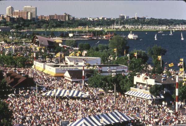 Bird's-eye view of a large crowd of music fans gathered at Summerfest, looking northeast toward the current site of Discovery World. Sailboats are on Lake Michigan.