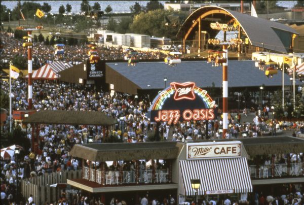Bird's-eye view of the Summerfest grounds. The Miller Jazz Oasis and Miller Cafe are in the foreground, and the TV 6 Comedy Cabaret and the Main Stage are in the background. A large crowd is in attendance, and people are riding the SkyGlider above the crowds.