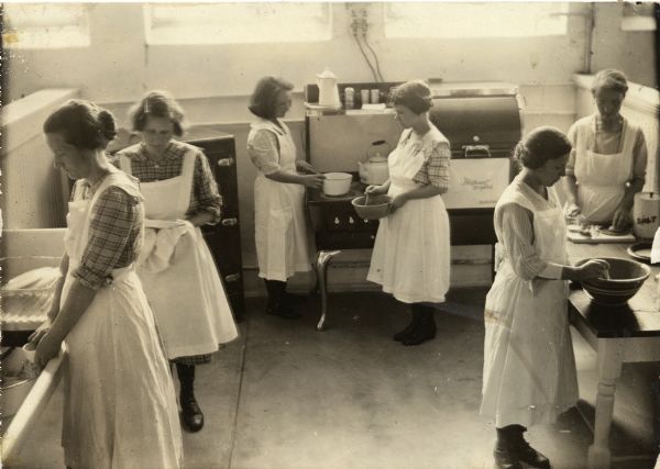 Slightly elevated view of six women wearing aprons learning domestic skills in a kitchen classroom. Two women on the left stand at a sink washing dishes, two women stand at a stove cooking in the center, and two prepare food at a table on the right.
