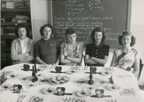 Five women are sitting at a table set for perhaps a Valentine's Day party. The setting was likely a Home Economics project for the women at the Northern Wisconsin Colony and Training School. There is a recipe written on a chalkboard behind the women.