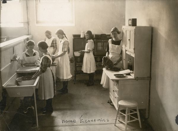 Slightly elevated view of six young women posed in a kitchen. Two women on the left stand at a sink washing dishes, two women stand at a table behind them, perhaps preparing food, one woman stands at a stove in the center, and one woman stands on the right with a mixing bowl. The women are residents at Northern Wisconsin Center for the Developmentally Disabled taking part in Home Economics training.