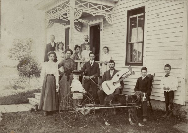Emory Bennett and family posed in front of the Bennett home. A young boy sits in a cart pulled by a dog. A seated man holds a harp guitar and another man standing beside him holds a violin.