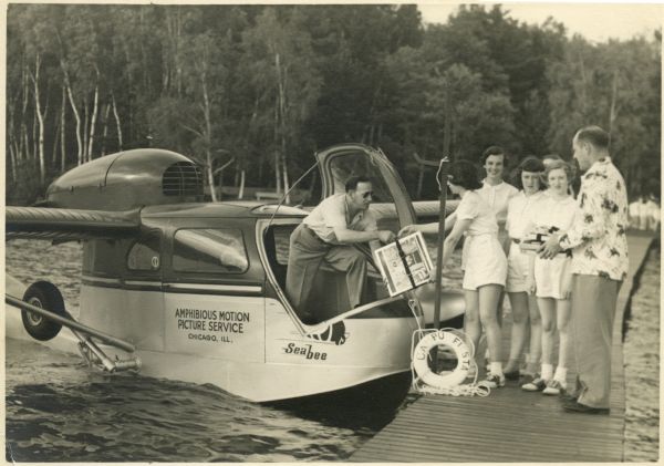 A man, possibly Benton C. Black, delivering films to Campo Fiesta at Trout Lake. The delivery is being made in a Seabee seaplane by the Amphibious Motion Picture Service of Chicago. A man, probably Warren Kirby, and five young women stand on a pier to meet the plane.