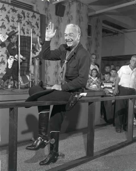 William Boyd as Hopalong Cassidy posing while sitting on a rail fence before meeting fans at Schuster's Department Store (N .12th and W. Vliet Streets) in Milwaukee. Several children are waiting in the background. In the background on the left a child mannequin is outfitted in cowboy shirt, pants, boots and a cowboy hat.