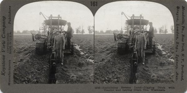 Stereograph showing workers digging a ditch with a tractor and laying drain tile.