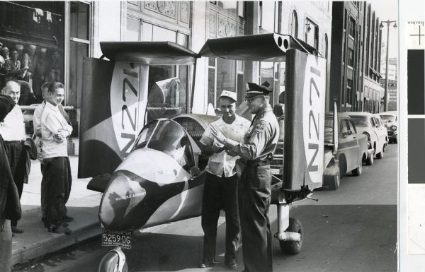 Milwaukee police Sergeant Edward Daily explains to Leland Bryan that his Roadable Aircraft, a combination airplane and automobile, may not be driven on Milwaukee's streets. A crowd watches from a nearby window and others gather on the sidewalk to see the unusual vehicle parked in front of the Milwaukee Journal building at 333 W. State Street.