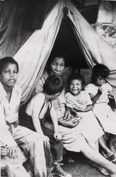 Woman and four children sing in front of a makeshift tent that is used as their home.